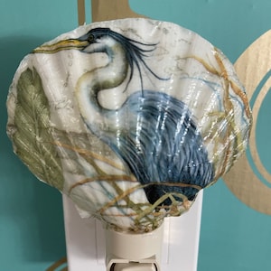May include: A night light shaped like a seashell with a blue heron printed on it. The night light is plugged into an electrical outlet.