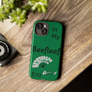 May include: Green phone case with the text "In My Beefleaf Era". The case features a fan with shamrock accents and a fishbone illustration. The phone case is on a wooden surface.