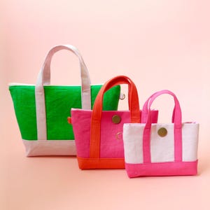 Può includere: Tre borse tote in tela di diverse dimensioni. La più grande è verde con dettagli bianchi, la media è rosa con dettagli arancioni e la più piccola è bianca con dettagli rosa. Ogni borsa ha un manico e una base.