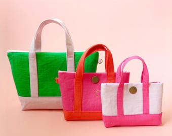 May include: Three colorful tote bags in varying sizes. The largest is green with white accents, the middle is pink with orange accents, and the smallest is white with pink accents. Each bag has a handle and a button closure.