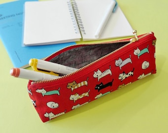 May include: A red fabric pencil case featuring a repeating pattern of small cats in various colors and poses. The case has a gold-toned metal zipper closure and contains two pens, one with a white barrel and one with a yellow barrel.