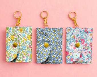 May include: Three small floral print keychains in yellow, blue, and pink. Each keychain has a gold-colored clasp and a snap closure. The keychains are displayed on a pink background.