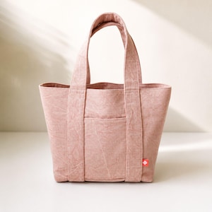 May include: A dusty rose tote bag with a textured fabric finish, featuring two sturdy top handles, a central exterior pocket, and a small rectangular red brand tag attached to the lower right side of the front panel of the bag.