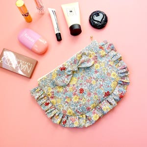 Könnte beinhalten: Eine Kosmetiktasche mit Blumenmuster, Rüschenkante und Schleifendetail. Die Tasche zeigt eine Mischung aus blauen, gelben und roten Blumen auf weißem Grund. Make-up-Produkte, darunter Lippenstift, Lidschatten und Lotion, sind um die Tasche herum angeordnet.