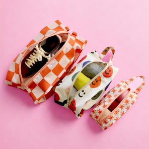May include: Three zippered travel bags in different patterns. One bag is orange and white checkered, another has colorful smiley faces, and the third has orange stars. The bags are open, revealing shoes and clothing.