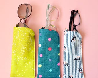 May include: Three fabric eyeglass cases standing upright. The left case is lime green with white speckles, the center case is teal with multicolored polka dots, and the right case features a blue and white striped pattern with French bulldog illustrations.