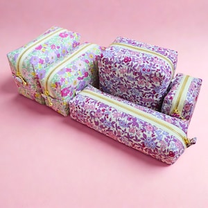 Puede incluir: Una colección de neceseres y estuches con estampado floral. Las bolsas presentan un fondo blanco con estampados florales rosas, morados y verdes. Cada bolsa tiene una cremallera dorada.