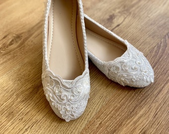 Op de afbeelding: Een paar ivoorkleurige ballerina's met ingewikkelde kanten details. De schoenen hebben een ronde neus en een delicate geschulpte rand. Ze liggen op een lichtgekleurde houten ondergrond, wat hun elegante ontwerp benadrukt.