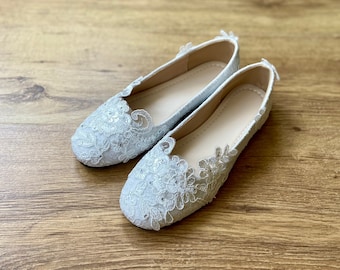 Op de afbeelding: Een paar lichtblauwe ballerina's met ingewikkelde kanten details en kleine versieringen. De schoenen hebben een ronde neus en een neutrale binnenzool. De ballerina's worden gepresenteerd op een houten oppervlak.