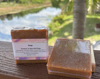 May include: Two bars of turmeric and kojic acid soap. The soap is a light brown colour and is wrapped in clear plastic. The soap is labelled "Soap, Turmeric & Kojic Acid Soap, Ingredients: Distilled Water, Saponified Oils of Olive, Coconut, Jojoba, Cocoa Butter, Shea Butter, Tea Tree & Lemongrass, Net Wt 3-5.02".
