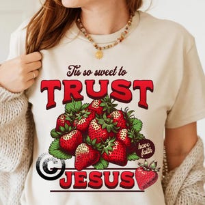 May include: Beige t-shirt featuring a graphic of strawberries and the words "Tis so sweet to TRUST JESUS" in red. The design includes a cluster of red strawberries with green leaves and a small strawberry on the bottom right.