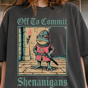 May include: A charcoal grey t-shirt featuring a graphic of a frog dressed as a medieval knight in pink armour with a sword. The text above reads Off To Commit and the text below reads Shenanigans. The frog stands on a stone floor near a torch.