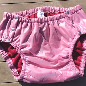 Könnte beinhalten: Rosa wasserdichte Windelhose mit Druckknöpfen. Die Innenseite ist mit rotem Fleece gefüttert. Der elastische Bund und die Beinöffnungen sind gerafft. Die Windelhose besteht aus glänzendem, glattem Material.