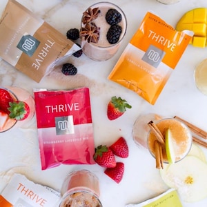 May include: An overhead shot features various Thrive lifestyle mix packets and prepared drinks. Flavors include chocolate, vanilla, strawberry, and cinnamon pumpkin spice. Fresh fruit and spices like strawberries, blackberries, and cinnamon sticks are scattered around the drinks.