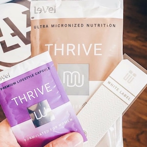 May include: A purple and white packet of Thrive Premium Lifestyle Capsules, formulated for women. The packet is labeled "Thrive" and "Lovel". The packet is sitting on top of a white and brown package labeled "Lovel Thrive Ultra Micronized Nutrition".