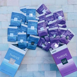 May include: A collection of Thrive premium lifestyle capsule packets in blue and purple. The blue packets are labeled formulated for men with an m logo, and the purple packets are labeled formulated for women with a w logo. Two larger boxes are at the base.