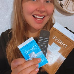 May include: A woman wearing a black baseball cap with the Adidas logo in white is holding three packets of Thrive nutritional supplements. The packets are blue, black, and brown. The brown packet has the text "THRIVE" in white.