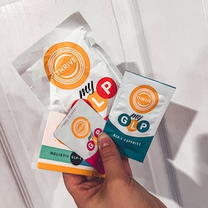 May include: Three small packages with the word "THRIVE" and the phrase "my GLP" printed on them. The packages are white with yellow, blue, and green accents. The text "HOLISTIC GLP-1" is printed on a card in the foreground.