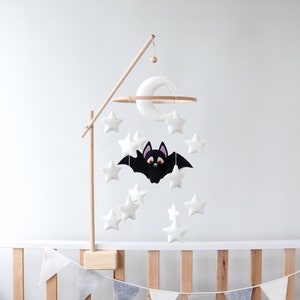 May include: A white felt baby mobile with a black felt bat, white felt stars, and a white felt crescent moon. The mobile is hanging from a wooden stand.