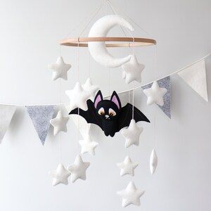 May include: A white felt moon and stars mobile with a black felt bat hanging from a wooden ring. The mobile is hanging from a white ceiling.