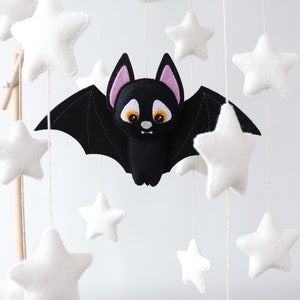 May include: A black felt bat with pink ears and orange eyes hangs from a wooden mobile with white felt stars and a crescent moon.