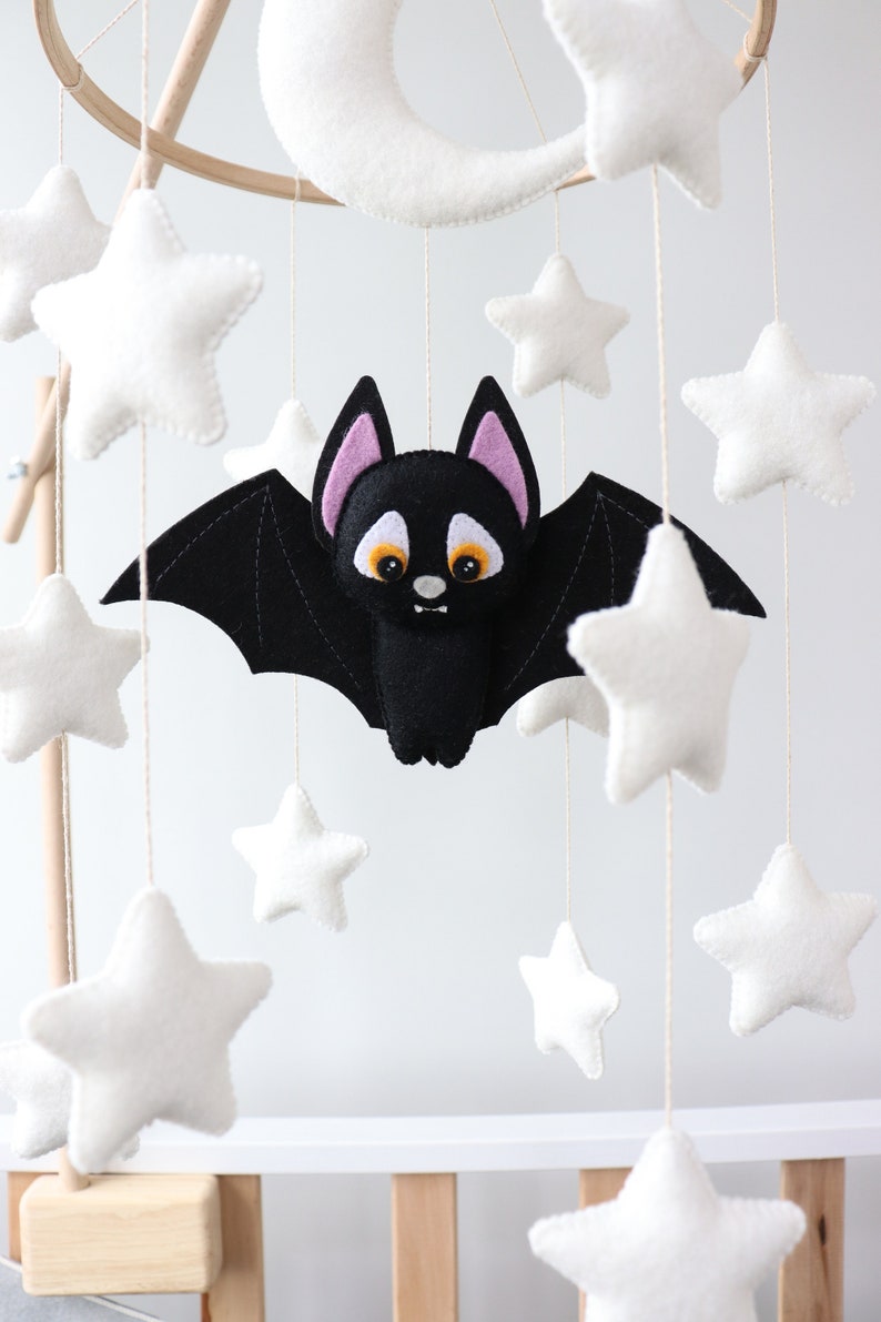 May include: A black felt bat with pink ears and orange eyes hangs from a wooden mobile with white felt stars and a crescent moon.