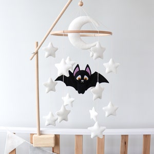 May include: A white felt moon and stars mobile with a black felt bat hanging from a wooden hoop. The mobile is attached to a wooden stand and is hanging over a crib with a gray and white felt banner.