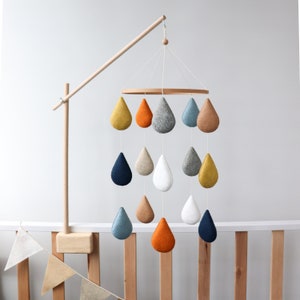 May include: A wooden mobile with a circular frame and multiple felt raindrop shapes in various colors, including blue, yellow, orange, and gray. The mobile is suspended from a wooden arm.