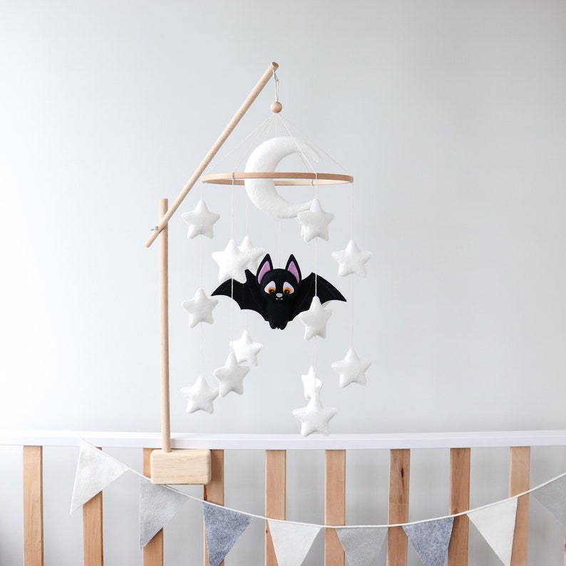 May include: A white felt baby mobile with a black felt bat, white felt stars, and a white felt crescent moon. The mobile is hanging from a wooden stand.