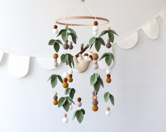 May include: A baby mobile featuring a sloth hanging from a felt tree branch, surrounded by felt leaves and hanging beads in shades of green, brown, and white. The mobile is suspended from a wooden ring and a white string.