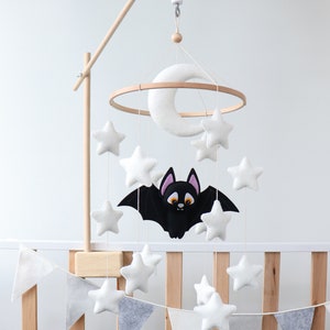 May include: A white and gray felt baby mobile with a black bat, moon, and stars hanging from a wooden ring. The mobile is attached to a wooden stand with a white music box.