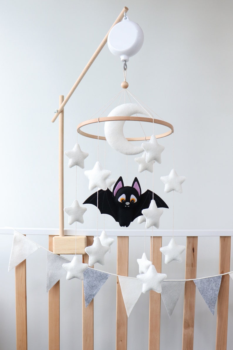 May include: A white and gray felt baby mobile with a black bat, moon, and stars hanging from a wooden ring. The mobile is attached to a wooden stand with a white music box.