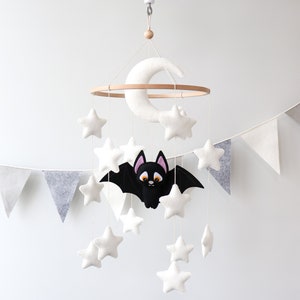 May include: A white felt baby mobile with a crescent moon, stars, and a black bat with orange eyes. The mobile is hanging from a white ceiling hook.