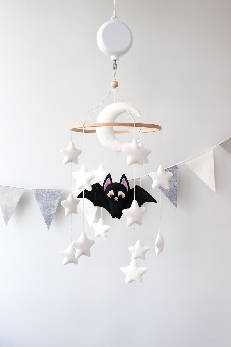 May include: A white felt baby mobile with a crescent moon, stars, and a black bat with orange eyes. The mobile is hanging from a white ceiling hook.