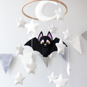 May include: A white felt crescent moon and stars hang from a wooden hoop with a black felt bat with orange eyes and pink ears in the center. The mobile is hanging from a white ceiling.