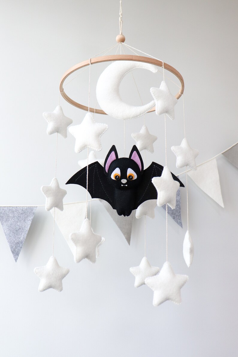 May include: A white felt crescent moon and stars hang from a wooden hoop with a black felt bat with orange eyes and pink ears in the center. The mobile is hanging from a white ceiling.