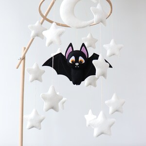 May include: A white felt crescent moon and stars hang from a wooden mobile with a black felt bat in the center. The mobile is attached to a wooden stand.