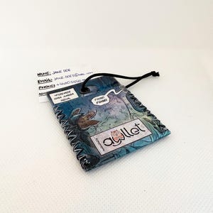 May include: A rectangular luggage tag featuring a comic book illustration of a creature, with speech bubbles containing the text FIZEMOS UMA AMIGA NOVA and FOGE! FOGE!, a logo with an owl and the word awllet, and a black elastic cord loop.