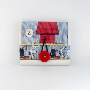 Op de afbeelding: Een kleine, rechthoekige portemonnee met een stripverhaalontwerp met Snoopy en zijn rode hondenhok. De portemonnee heeft een rode knoopsluiting en een zwart touwtje. De achtergrond is lichtblauw.