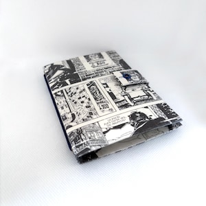 Könnte beinhalten: Ein kleines, gefaltetes Portemonnaie mit einem schwarz-weißen Comic-Print. Das Portemonnaie hat einen dunkelblauen Rand und einen blauen Knopfverschluss. Die Comic-Panels sind auf der Außenseite mit Text und Illustrationen sichtbar.