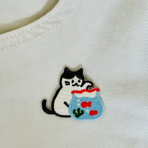 May include: A white and black cat with a black nose and eyes is holding a blue fishbowl with two red fish and a green plant inside. The cat is looking at the fishbowl.