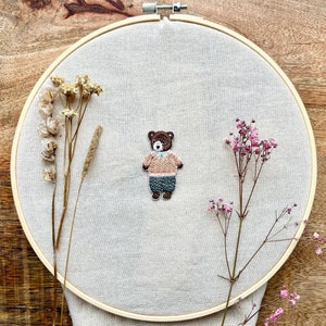 May include: A brown bear embroidered on a beige fabric stretched over a wooden embroidery hoop. The bear is wearing a pink shirt and blue trousers. Dried flowers are arranged around the hoop.