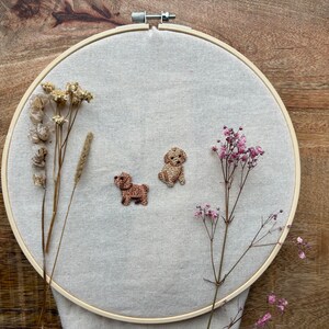 May include: Two embroidered brown and beige dogs on a beige fabric stretched over a wooden embroidery hoop. Dried flowers are on either side of the dogs.