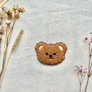 May include: A brown embroidered bear face patch with a fluffy texture. The patch is on a beige fabric background.