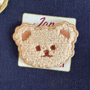 May include: A brown and white fuzzy bear face embroidered patch. The bear has brown eyes and a brown nose.