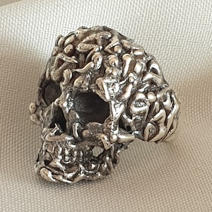 Puede incluir: Un anillo de calavera de plata con un diseño detallado e intrincado. La calavera está cubierta con un patrón de pequeños detalles en relieve que se asemejan a hojas o vides.
