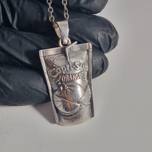 Puede incluir: Collar con colgante de plata con diseño de bolsa de zumo Capri-Sun. El colgante tiene las palabras "Capri Sun ORANGE" grabadas, junto con una ilustración de naranja. El collar tiene una cadena de plata.
