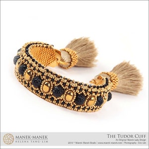 Peut inclure: Un bracelet en perles dor&eacute;es et noires avec des accents de pierres pr&eacute;cieuses noires et deux pompons beiges. Le bracelet s'appelle "The Tudor Cuff" et est un design original de Manek Lady.