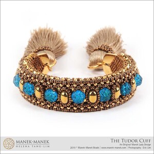 Peut inclure: Un bracelet en perles dor&eacute;es et brunes avec des pierres turquoise et des glands. Le bracelet s'appelle "The Tudor Cuff" et est un design original de Manek Lady.