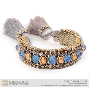 Peut inclure: Un bracelet en perles dor&eacute;es et argent&eacute;es avec des perles de verre bleues et des glands gris. Le bracelet s'appelle "The Tudor Cuff" et est un design original de Manek Lady.